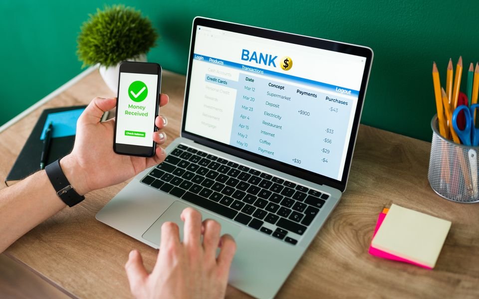 A person checking a bank account on a laptop while a phone confirms money received